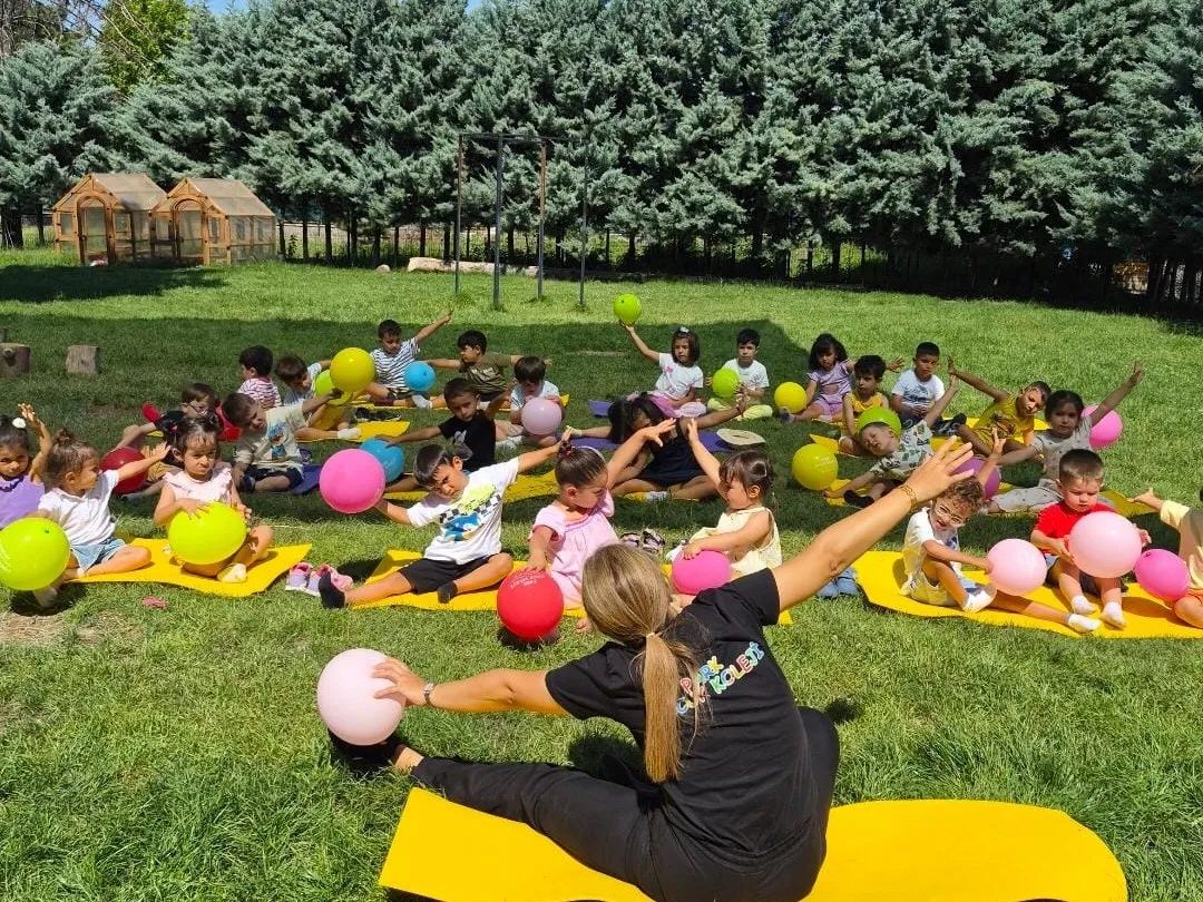 Bahçede minderler üzerinde balonlarla hareket çalışması yapan çocuklar ve öğretmenleri